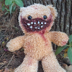 May include: A tan teddy bear with a gruesome, open-mouthed design. The bear has black button eyes, a blood-red mouth filled with white teeth, and red accents on its fur. The plush toy is set against a natural outdoor backdrop.