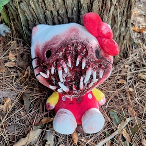 May include: A plush toy resembling a white cat character with a red bow, featuring a modified face with a large, open, blood-stained mouth filled with numerous sharp, white, pointed teeth. The body is red with yellow sleeves and white feet.