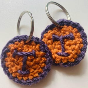 May include: Two handmade crochet keychains. Each keychain features a circular design with an orange center and a purple border. A purple letter "T" is crocheted in the center of each keychain. Silver key rings are attached to the top of each keychain.