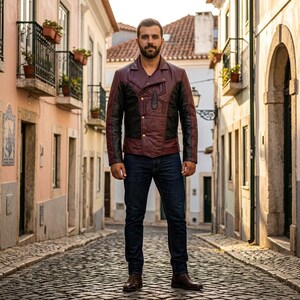 May include: A burgundy and black leather jacket with a button-up front and a unique spider emblem on the chest. The jacket has black sleeves and a burgundy body. The person is wearing dark blue jeans and brown boots.