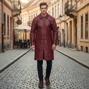 May include: A burgundy leather coat with a zip-up front, a collar, and a long length. The coat has a tailored fit and is worn with black pants and brown shoes. The background is a cobblestone street lined with buildings.