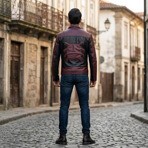 May include: A burgundy and black leather jacket with a stylized design on the back. The jacket has a classic collar and a front zipper. The person is wearing blue jeans and black boots. The background is a cobblestone street.