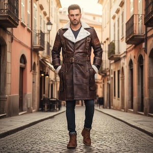 May include: A brown leather coat with a white shearling collar and cuffs. The coat features a belted waist, multiple pockets, and decorative buckles. The person is wearing dark blue jeans and brown boots. The background is a cobblestone street lined with buildings.