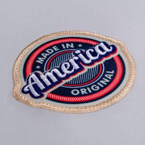 Puede incluir: Un parche bordado de forma ovalada con un centro azul oscuro y un borde circular rojo y blanco. El texto "MADE IN America ORIGINAL" está cosido con hilo blanco y azul en el centro, rodeado por un borde cosido de color beige.