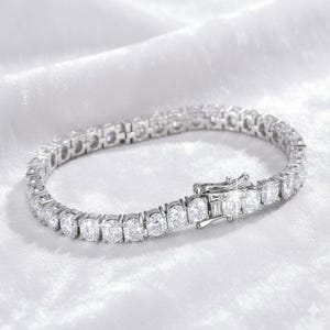 May include: A silver tennis bracelet with numerous round, clear gemstones. The bracelet is set on a white, textured surface. The clasp is visible and adds to the overall sparkle of the jewelry.