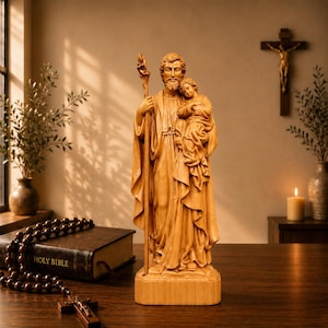 Op de afbeelding: Een lichtbruin houten beeld van Sint-Jozef met een kind, met een staf. Het beeld staat op een houten voet. Een bijbel en rozenkrans liggen op de voorgrond. Een kruisbeeld staat op de achtergrond.