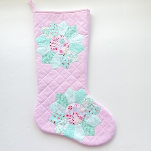 May include: Pink quilted Christmas stocking with two large, decorative flower appliqués. The flowers feature a mix of light blue and patterned fabrics with candy and snowman motifs. The stocking has a gingham pattern and a hanging loop.