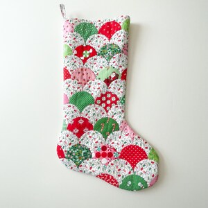 Puede incluir: Un calcetín navideño hecho a mano con un diseño de vieira. El calcetín presenta un patrón de patchwork de formas de abanico rojas, verdes y rosas, cada una con un estampado festivo diferente. El fondo es blanco con una guirnalda de luces.