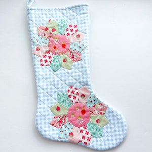 May include: A quilted Christmas stocking with a light blue and white gingham pattern. The stocking features two large floral designs with pink, red, and green strawberry and floral fabric appliqués. A white loop is attached at the top.