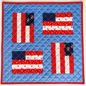 May include: A quilted square featuring four stylized American flags. Each flag has red and white stripes, a blue field with white dots, and a red border. The background is blue with white polka dots, and the edges are trimmed with a red border.