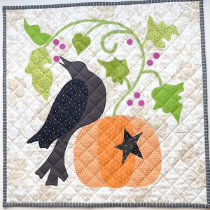 May include: A quilted wall hanging featuring a black crow perched on an orange pumpkin with a black star. Green leaves and purple berries adorn the vines. The background is a floral pattern with a gray border.