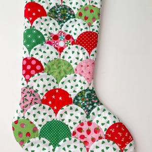 Puede incluir: Un calcetín navideño acolchado con un diseño festoneado. El calcetín presenta varias telas con patrones rojos, verdes y rosas, incluyendo lunares, estrellas, fresas e estampados florales, sobre un fondo blanco con pequeños árboles de Navidad verdes.