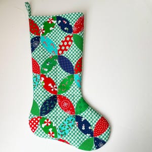 May include: A green Christmas stocking with a patchwork design featuring red, blue, and green fabric with various holiday patterns. The stocking is made of quilted fabric and has a white loop at the top.
