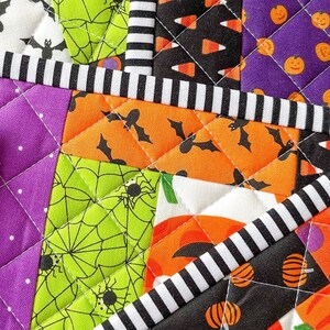 May include: A patchwork quilt with a Halloween theme. The quilt is made of various fabrics with different patterns, including bats, spiders, candy corn, and pumpkins. The quilt is sewn together with white thread and has a black and white striped border.