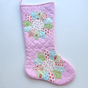 May include: A pink quilted Christmas stocking with two patchwork flower designs in shades of green, blue, and pink. The stocking has a white background with a red and white candy cane pattern.
