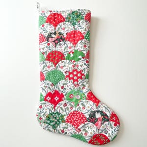 May include: A Christmas stocking with a patchwork design. The stocking features a variety of red and green patterned fabrics, including polka dots, floral prints, and Christmas-themed motifs. The stocking is approximately 20 inches long and has a loop for hanging.