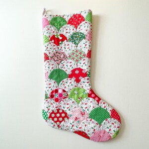 May include: A handmade Christmas stocking with a scalloped design. The stocking features red, green, and pink fabric circles with various patterns, including polka dots, snowflakes, and Christmas lights, on a white background.