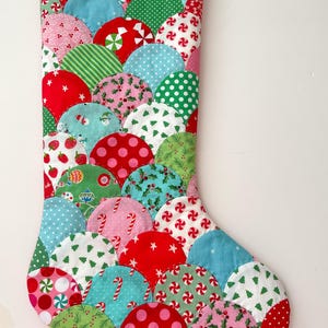 Puede incluir: Un calcetín navideño hecho a mano con un diseño de patchwork. El calcetín presenta una variedad de telas en rojo, verde, azul y rosa, con patrones que incluyen lunares, rayas, bastones de caramelo y árboles de Navidad. Tiene un lazo rojo para colgar.