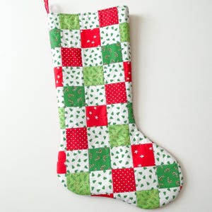 May include: A quilted Christmas stocking with a patchwork design. The squares are red, green, and white, featuring patterns of stars, Christmas trees, and candy canes. A red ribbon loop is attached at the top for hanging.