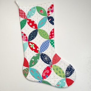 May include: A white Christmas stocking with a quilted design featuring red, green, blue, and pink fabric circles with various holiday patterns.