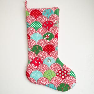 Puede incluir: Una media de Navidad roja, verde y azul con un patrón acolchado de conchas superpuestas. La media está decorada con una variedad de telas con temática navideña, incluyendo bastones de caramelo, copos de nieve y árboles de Navidad.