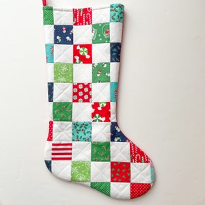 May include: A white Christmas stocking with a patchwork design featuring red, green, blue, and white squares. Each square has a different holiday-themed pattern, including candy canes, snowmen, and holly.