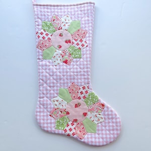 May include: Pink and white gingham Christmas stocking with two floral appliqués. The appliqués feature pink, green, and white patterned fabrics with strawberry and swirl designs. The stocking is quilted.