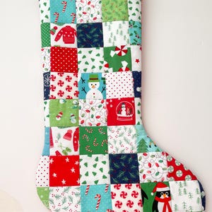 May include: A quilted Christmas stocking with a patchwork design. The squares feature holiday-themed patterns like snowmen, candy canes, and Christmas trees in red, green, blue, and white. A red loop is at the top for hanging.