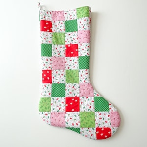 May include: A quilted Christmas stocking with a patchwork design. The squares are red, green, and pink, with patterns of stars, polka dots, and Christmas lights. The stocking has a loop for hanging.