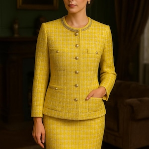 Op de afbeelding: Een gele tweed rokpak. De jas heeft gouden knopen en zakdetails, met een decoratieve kraag met kralen. Het pak is een klassieke stijl, geschikt voor professionele of formele gelegenheden. De stof is geweven.