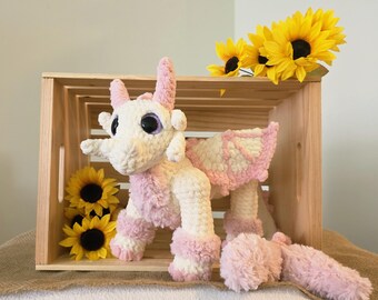 May include: A handmade, plush dragon toy in a wooden crate. The dragon is white with pink accents on its wings, legs, and tail. The dragon has large, purple-rimmed eyes. Yellow sunflowers are placed around the dragon.