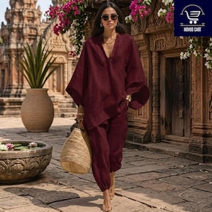 Op de afbeelding: Een diepbordeauxrode tweedelige set bestaande uit een tuniek met lange mouwen en een V-hals en een bijpassende losvallende broek. De outfit wordt gecombineerd met een grote, ronde, gevlochten strooien handtas. De stof heeft een zichtbare textuur.