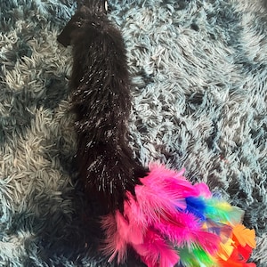 May include: A black faux fur tail with a vibrant, rainbow-colored feather accent. The feathers transition from pink to orange, with shades of blue, green, and yellow. The tail is designed to be worn as a costume accessory.