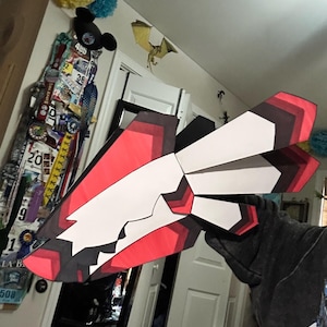 May include: A geometric, angular sculpture with a red, white, and black color scheme. The artwork appears to be made of layered paper or cardboard, with sharp angles and a stylized design. The piece is held up in front of a wall.