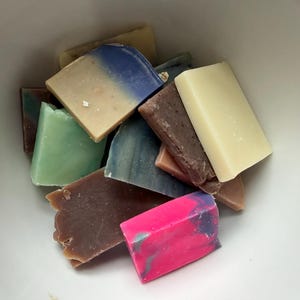Puede incluir: Una colección de jabones rectangulares coloridos en un cuenco blanco. Los jabones son de varios colores, incluyendo azul, verde, marrón, rosa y crema. Algunas barras tienen un efecto veteado. Los jabones están apilados juntos, llenando el cuenco.