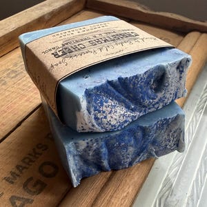 May include: Two bars of blue soap with a textured surface and a brown paper label. The label reads "Christmas Cheer" and "Hand & Body Cold Process Soap". The soap is stacked on a wooden surface.