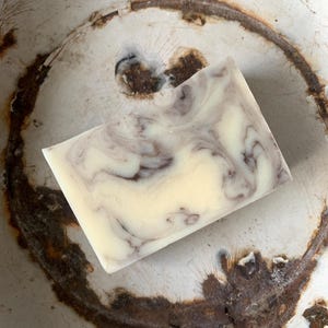 Op de afbeelding: Een rechthoekige blok handgemaakte zeep met een crèmewitte basis en wervelende donkerpaarse patronen. De zeep ligt op een verweerd, gebroken witte ondergrond met roestbruine markeringen, wat een rustieke uitstraling creëert.