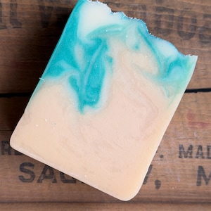 May include: A rectangular bar of soap featuring a marbled design with a creamy beige base color and swirling patterns of vibrant turquoise blue. The top edge of the soap bar has a slightly uneven, textured finish with a visible gradient effect.