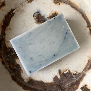 Peut inclure: Un pain de savon rectangulaire bleu clair avec des mouchetures foncées repose dans un bol blanc vieilli avec des taches de rouille brunes. Le savon a un aspect marbré, suggérant des ingrédients naturels. La texture du bol ajoute une touche rustique et vintage.