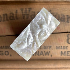 May include: A rectangular bar of soap with a pearlescent, swirling pattern. The soap is a creamy white colour with subtle iridescent highlights. The soap is angled against a wooden surface with faded text.