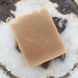 Puede incluir: Una pastilla rectangular de jabón marrón claro con una superficie texturizada. El jabón tiene un borde superior ligeramente irregular y está sobre un fondo blanco con un patrón circular oxidado.