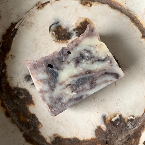 May include: A rectangular bar of handmade soap with a marbled appearance, featuring shades of purple, white, and gray. The soap is placed on a white, circular surface with a rusty brown ring around the edge. The soap has a slightly rough, textured surface.