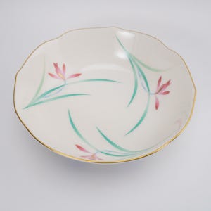 May include: A shallow, round, white porcelain bowl with a scalloped edge and a thin gold rim. The interior features a delicate painted design of pink flowers with long, slender green leaves arranged in a swirling pattern across the surface.