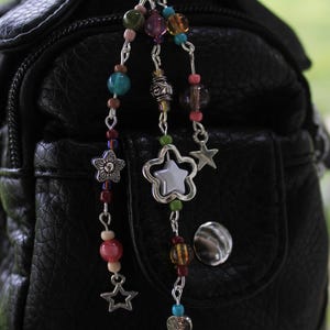 May include: A beaded charm with a variety of colorful beads, including pink, blue, green, and brown, hangs from a silver clasp. The charm features star and flower-shaped pendants. The charm is attached to a black leather bag.