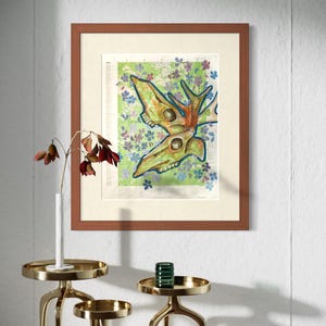 May include: Framed artwork featuring a stylized animal skull with antlers, painted in shades of green and orange, set against a floral background. The artwork is displayed in a brown wooden frame. Decorative gold side tables with a vase and a green glass candle holder are in the foreground.