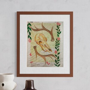 May include: Framed art print featuring a deer skull and antlers over a vintage map background. The artwork is accented with pink flowers and green leaves, all within a brown wooden frame. A unique piece for home decor.