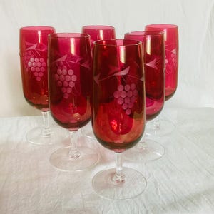 May include: A set of six ruby red glass goblets, each featuring a clear etched design of grape clusters and vine leaves. These stemmed glasses have a rounded bowl shape and a transparent base, showcasing a deep crimson color throughout the glass.
