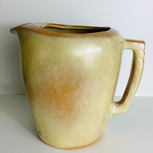 May include: A ceramic pitcher featuring a smooth, matte finish in shades of light yellow and tan. The vessel has a wide, open spout, a curved body, and a sturdy, angular handle attached to the side. The glaze shows subtle color variations.