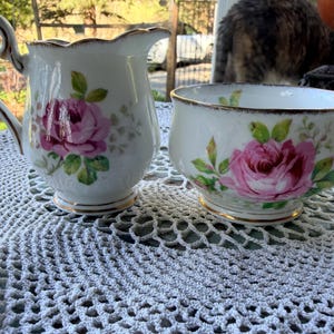 May include: A white porcelain creamer and sugar bowl set. Each piece is decorated with a large pink rose and green leaves. The rims and handles are trimmed with gold. The creamer has a curved handle and a spout.