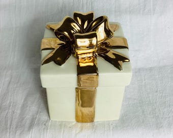 May include: A square ceramic box shaped like a gift, featuring a smooth off-white finish on the base and lid. The top of the lid is adorned with a large, reflective gold-colored ceramic bow, with gold ribbon accents extending down the sides.